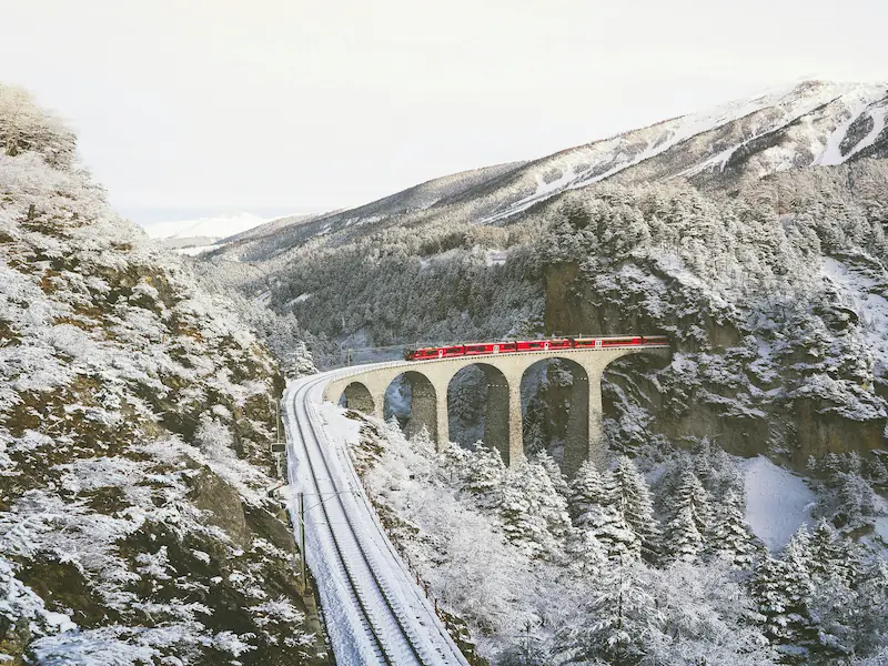 Red Rhaetian Railway train crossing a snow-covered stone viaduct in the Swiss Alps