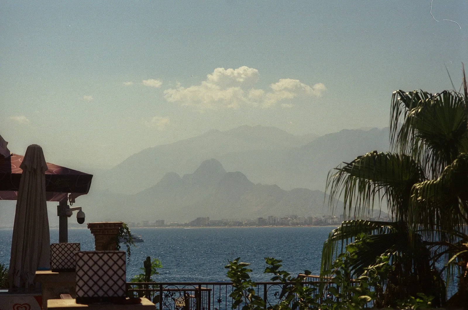 Hazy Taurus Mountains rising behind Antalya's coastline, viewed from a seaside terrace with palm fronds in the foreground