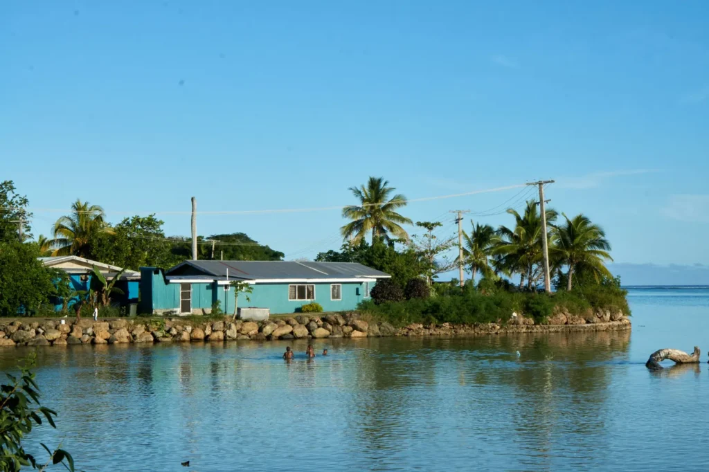 Children swimming in a calm lagoon beside a teal village house with palm trees in Fiji